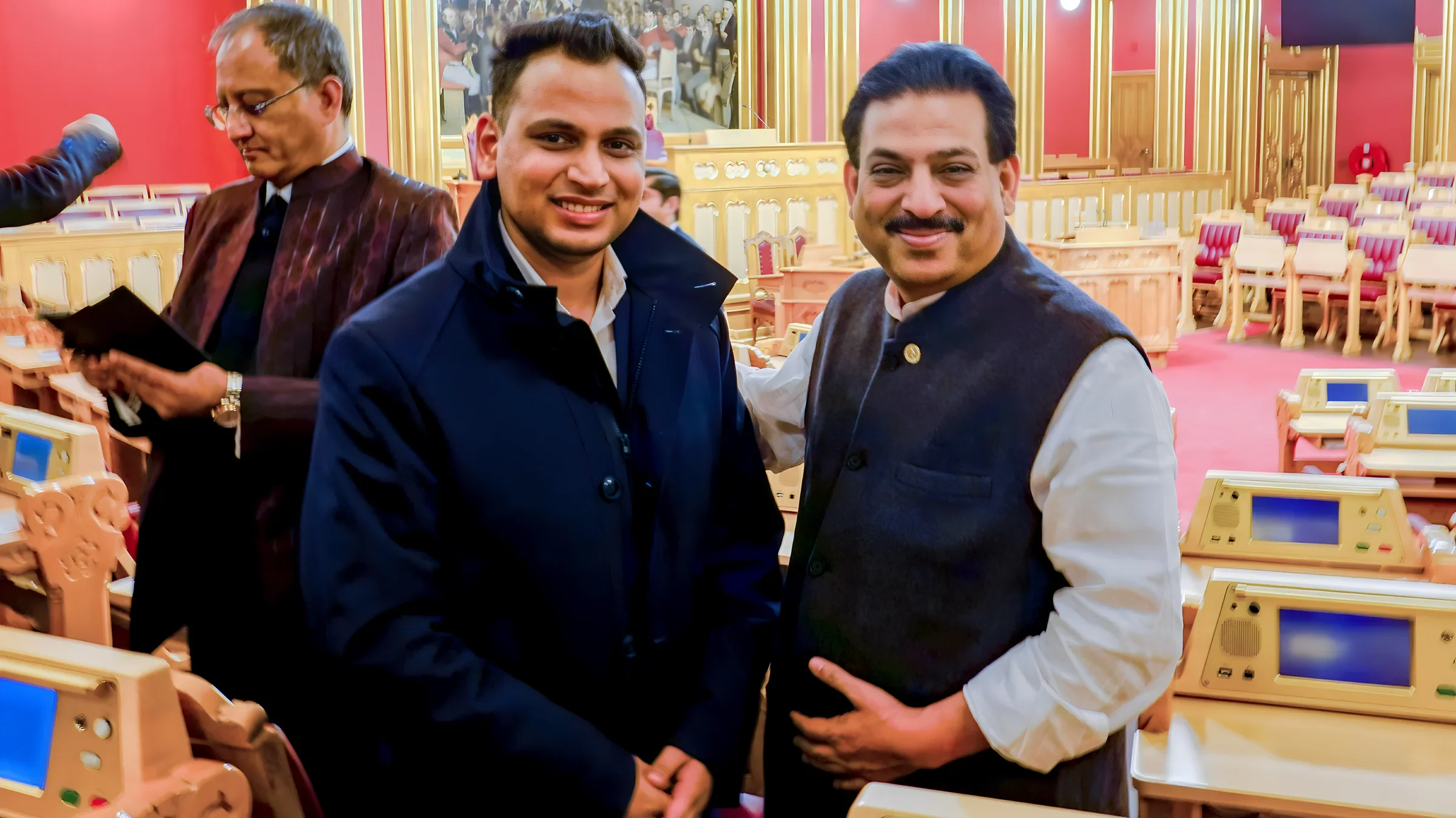 Lithvik Mukesh Sharma is photographed inside a formal legislative assembly hall alongside Vibhav Kant Upadhya. The hall features deep red/burgundy walls, ornate golden vertical paneling